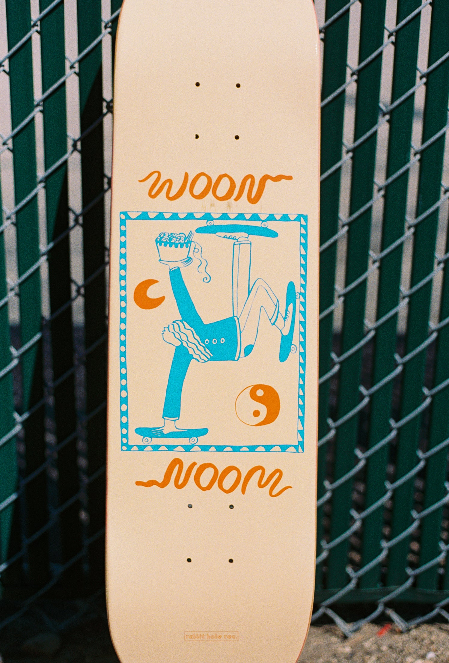 Skateboard with woon logo and person skating and eating noodles on it.