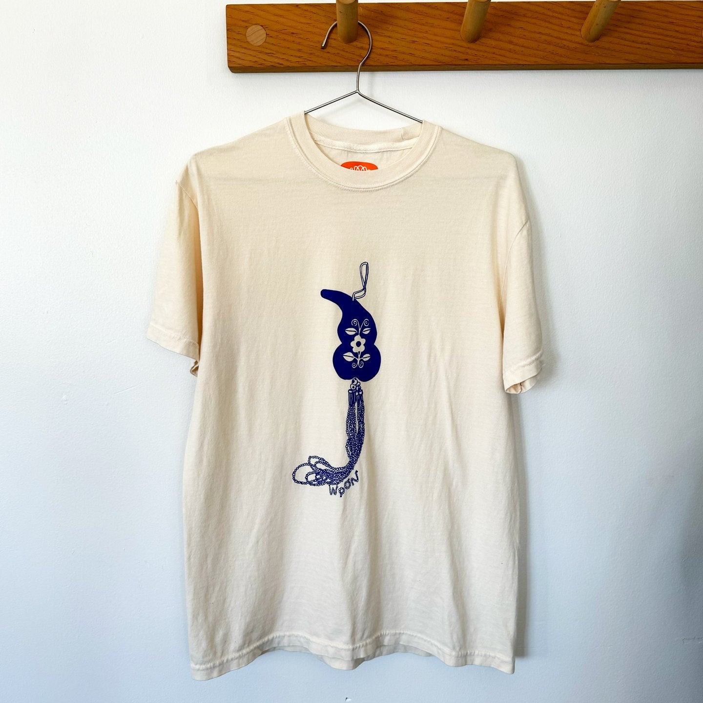 Cream color tee shirt hanging on a wooden hood. Picture on shirt is off a blue gourd key chain