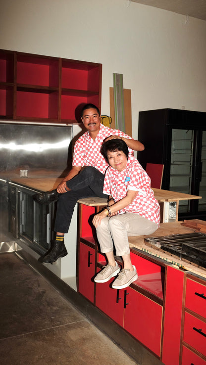 Two people wearing red checkered work shirts sitting on a work bench