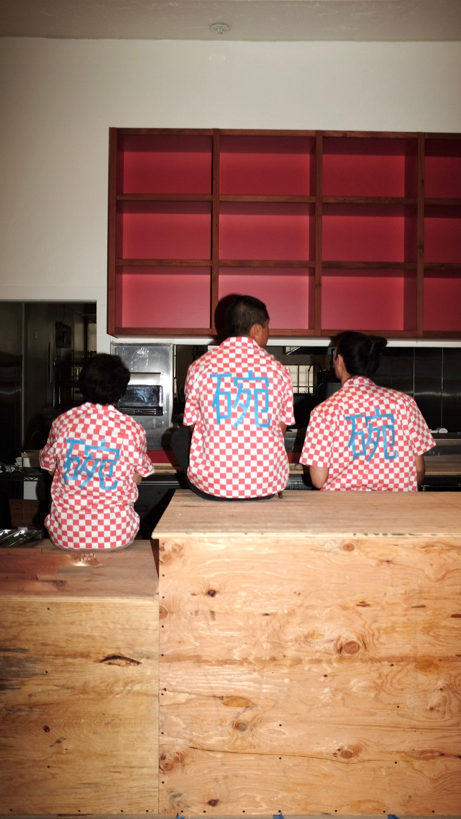 Three people sitting on a work bench wearing red checkered work shirts