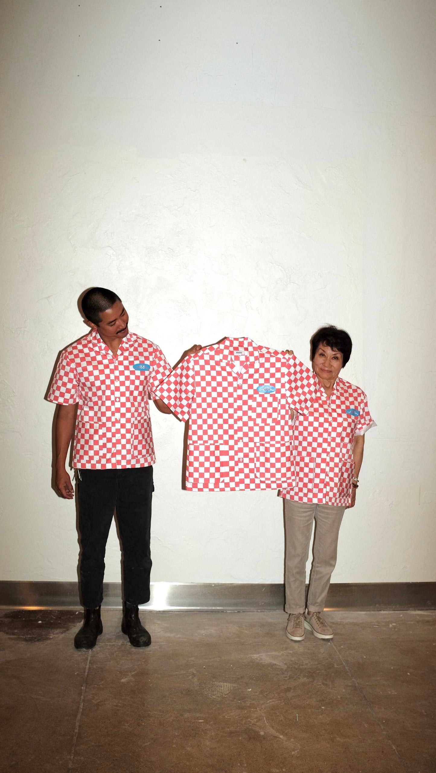 Two people wearing red checkered work shirts holding another red checkered work shirt showing the front