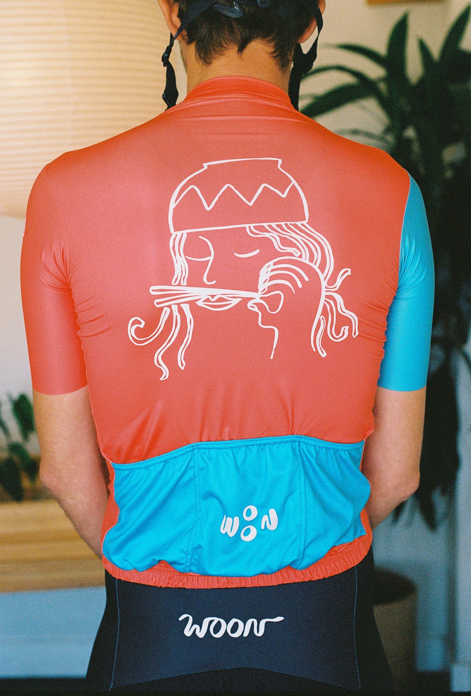 A close up of the back of a person wearing a cycling jersey. Cycling jersey has a person with a bowl of noodles dumped on his head and chopsticks for a mustache