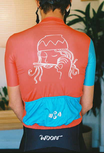 A close up of the back of a person wearing a cycling jersey. Cycling jersey has a person with a bowl of noodles dumped on his head and chopsticks for a mustache